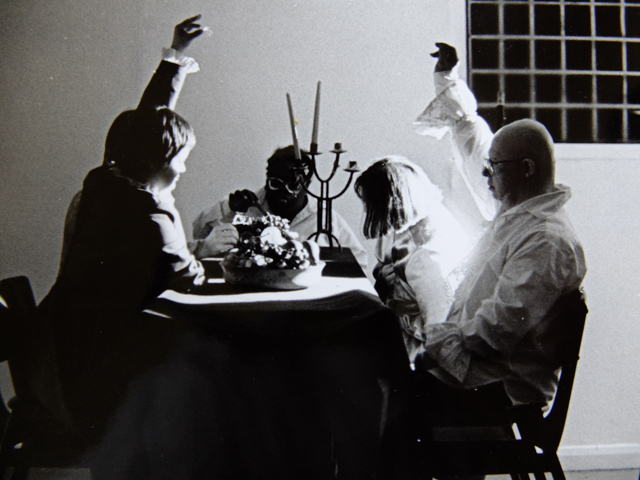 A black & white photo still of the performance Diary of a Dead Artist  by Corali Dance Company. The photo is taken in the original gallery of Southwark Park Galleries and features Laura Hayman, Natalie Jefferies, Margaret Courtman and Ray Walker. They sit at a table with a candlestick and fruit raised arms hight as if ready to take the fruit. The costumes are frilly blouses.